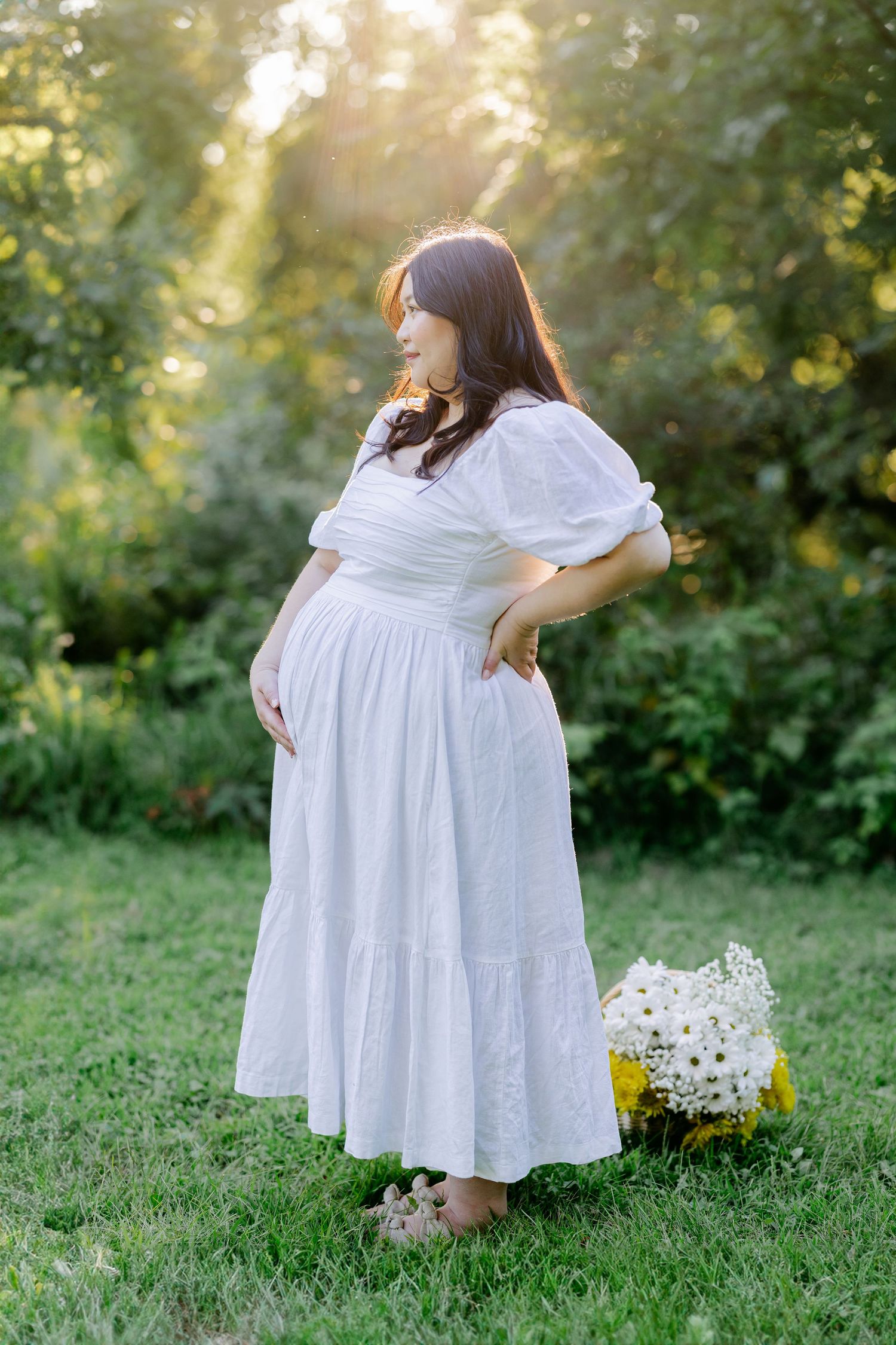 A pregnant woman in a flowing white dress stands in a garden at sunset with a bouquet of wildflowers nearby.