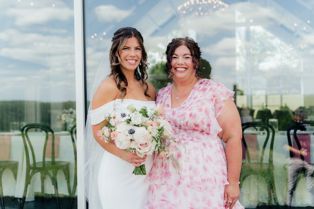 Two people stand together at a wedding venue, one in a white wedding dress and one in a pink floral dress.