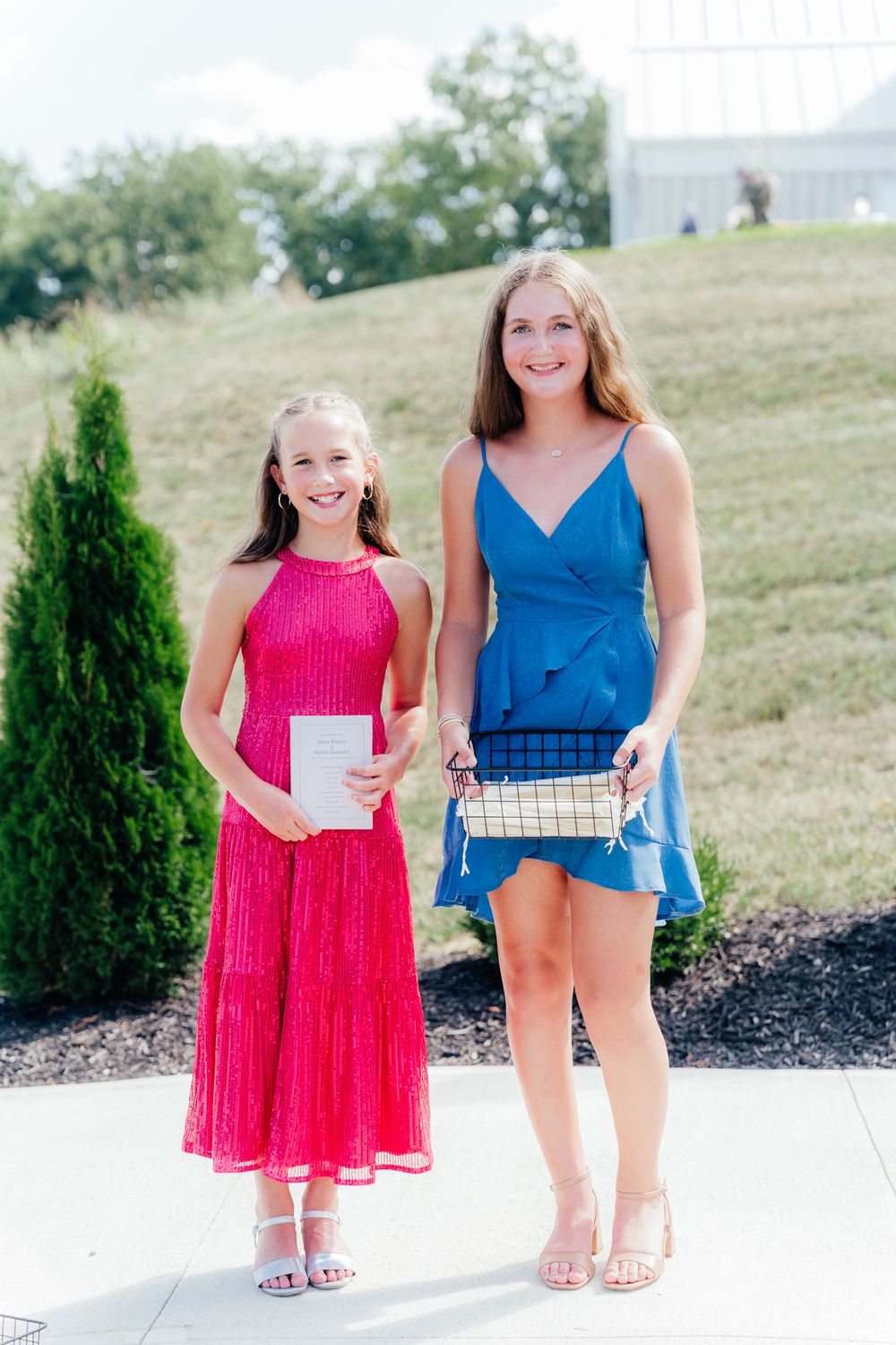 Two people in summer outfits stand outdoors, one wearing a blue dress and the other in a bright pink formal dress.