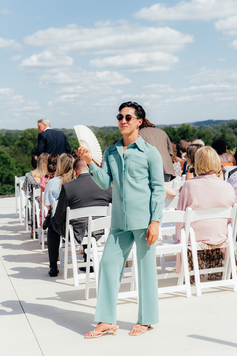 Someone in a mint green pantsuit and sunglasses stands among white folding chairs at an outdoor event venue.