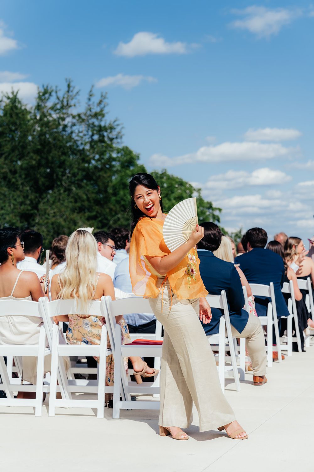 Event attendee holding a decorative paper fan at an outdoor wedding ceremony with white chairs and sunny blue skies.
