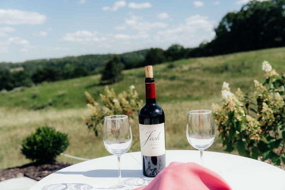Red wine bottle and two empty wine glasses sit on outdoor table with scenic countryside view in background.