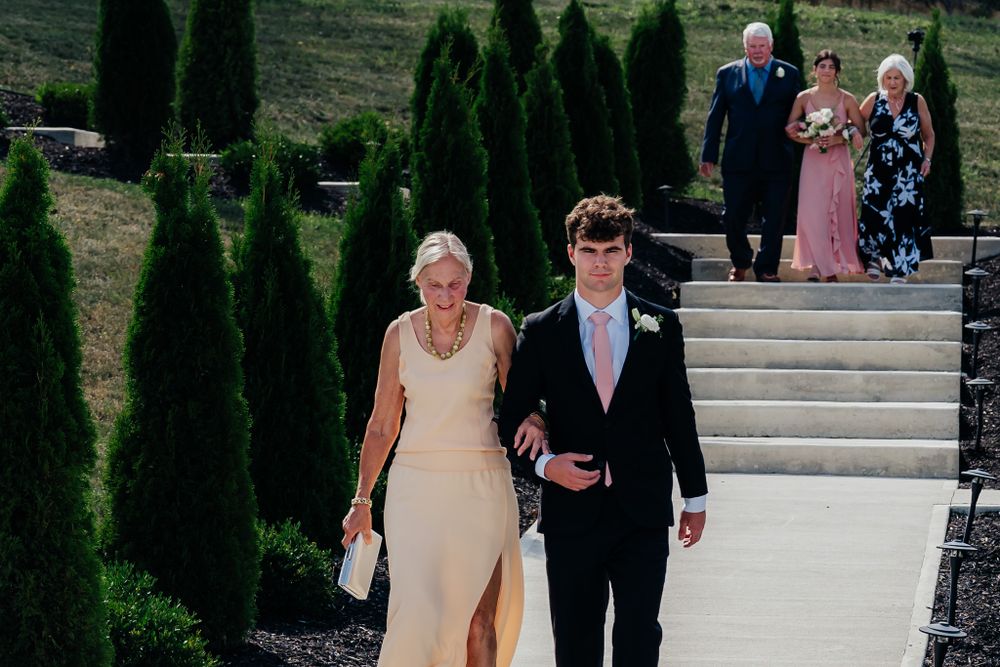 Well-dressed couple walks down stone steps at outdoor venue with cypress trees and guests in background.