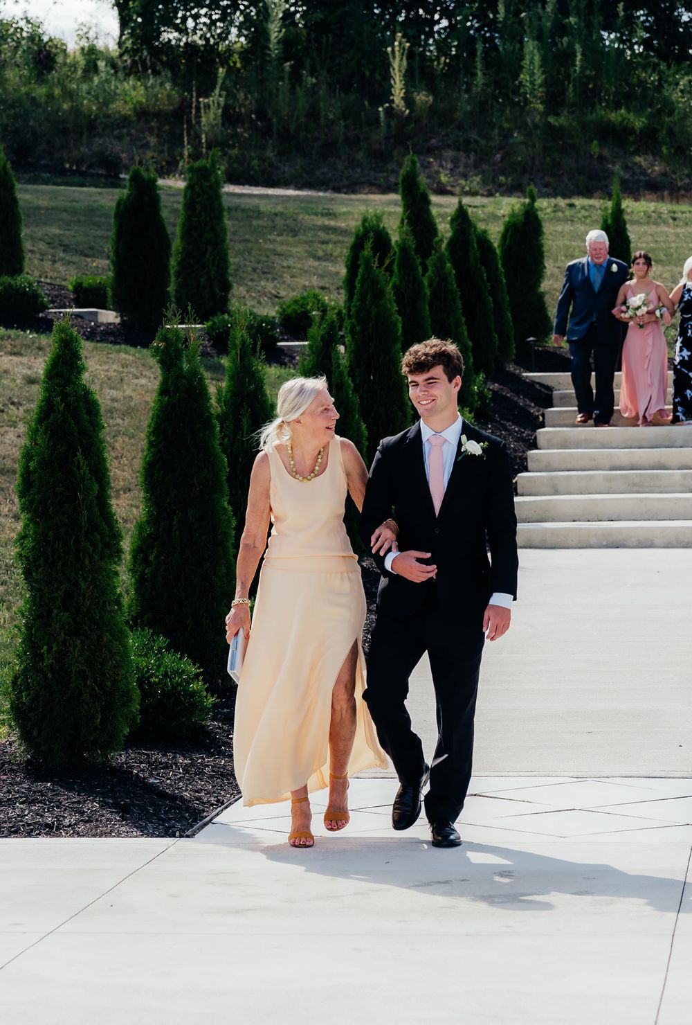 Couple walks down garden steps lined with tall green cypress trees on a sunny day.