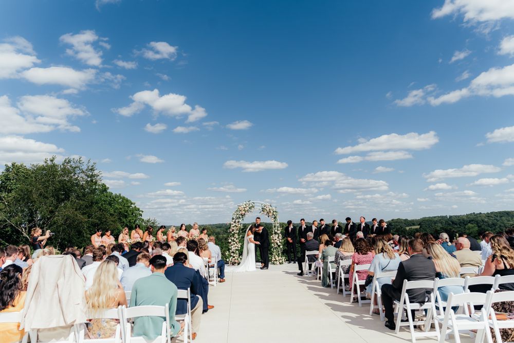 Outdoor wedding ceremony setup with white chairs and aisle on sunny day with blue sky and fluffy clouds.