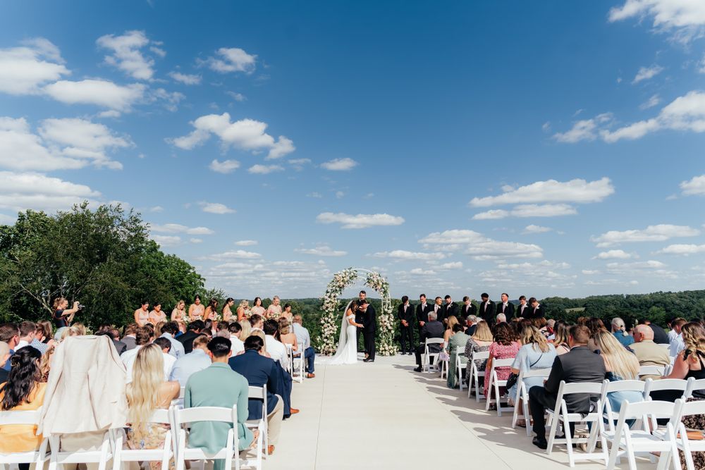 An outdoor wedding ceremony on a sunny day with guests seated in white chairs along a path against a blue sky backdrop.