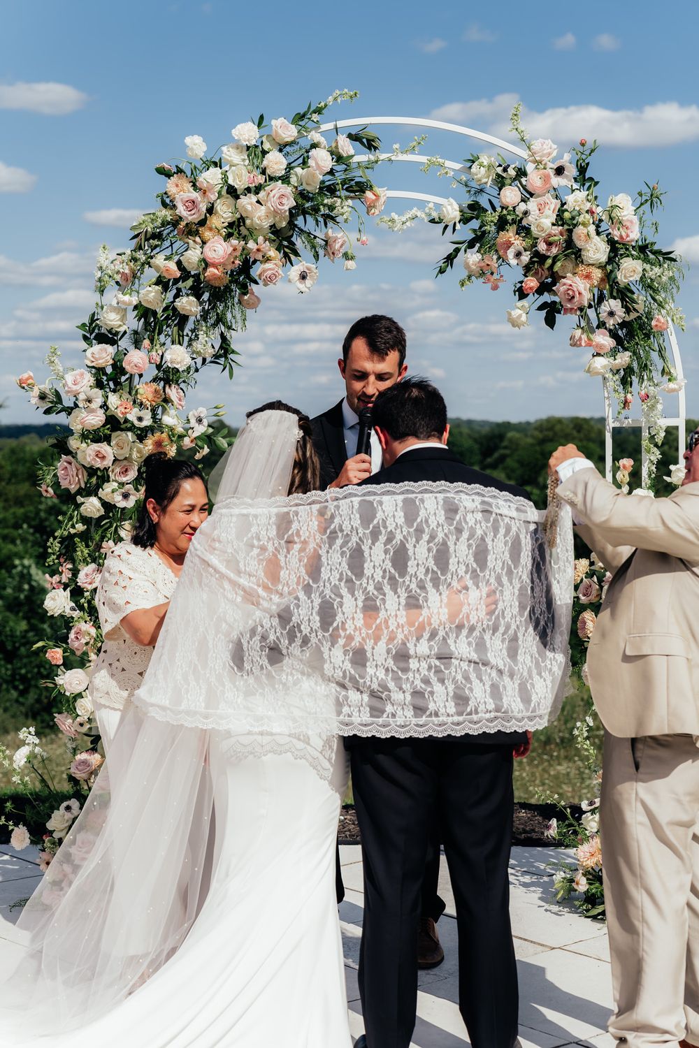 A wedding ceremony takes place under a beautiful floral arch with pink and white roses against a blue sky.