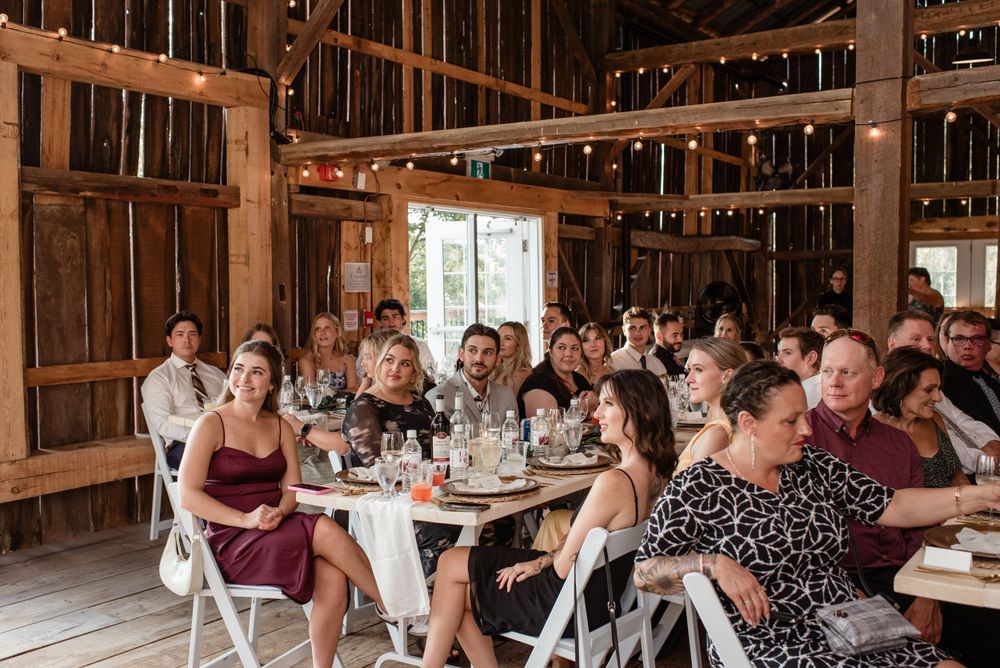 Wedding reception guests gather at tables inside a rustic wooden barn setting with natural light streaming through windows.