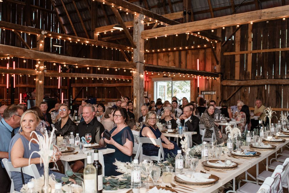 Long banquet tables set for a wedding reception in a barn with string lights and white table settings.