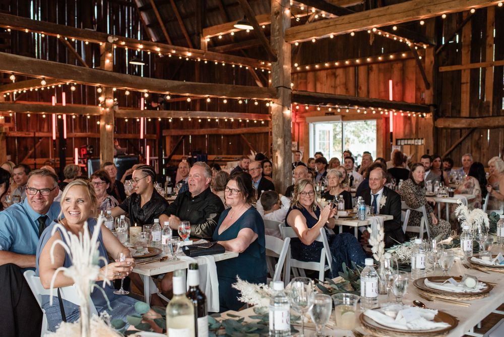 A large group of guests seated at elegantly set tables during a wedding reception in a rustic barn venue with string lights.