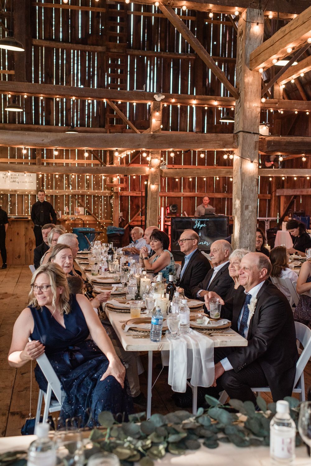 Wedding guests enjoy dinner at rustic barn reception with wooden beams and string lights creating warm ambiance.