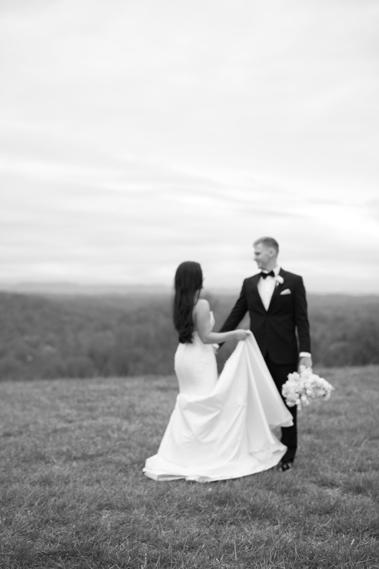 A bride and groom share a romantic moment on a hilltop overlooking a scenic valley landscape in black and white.