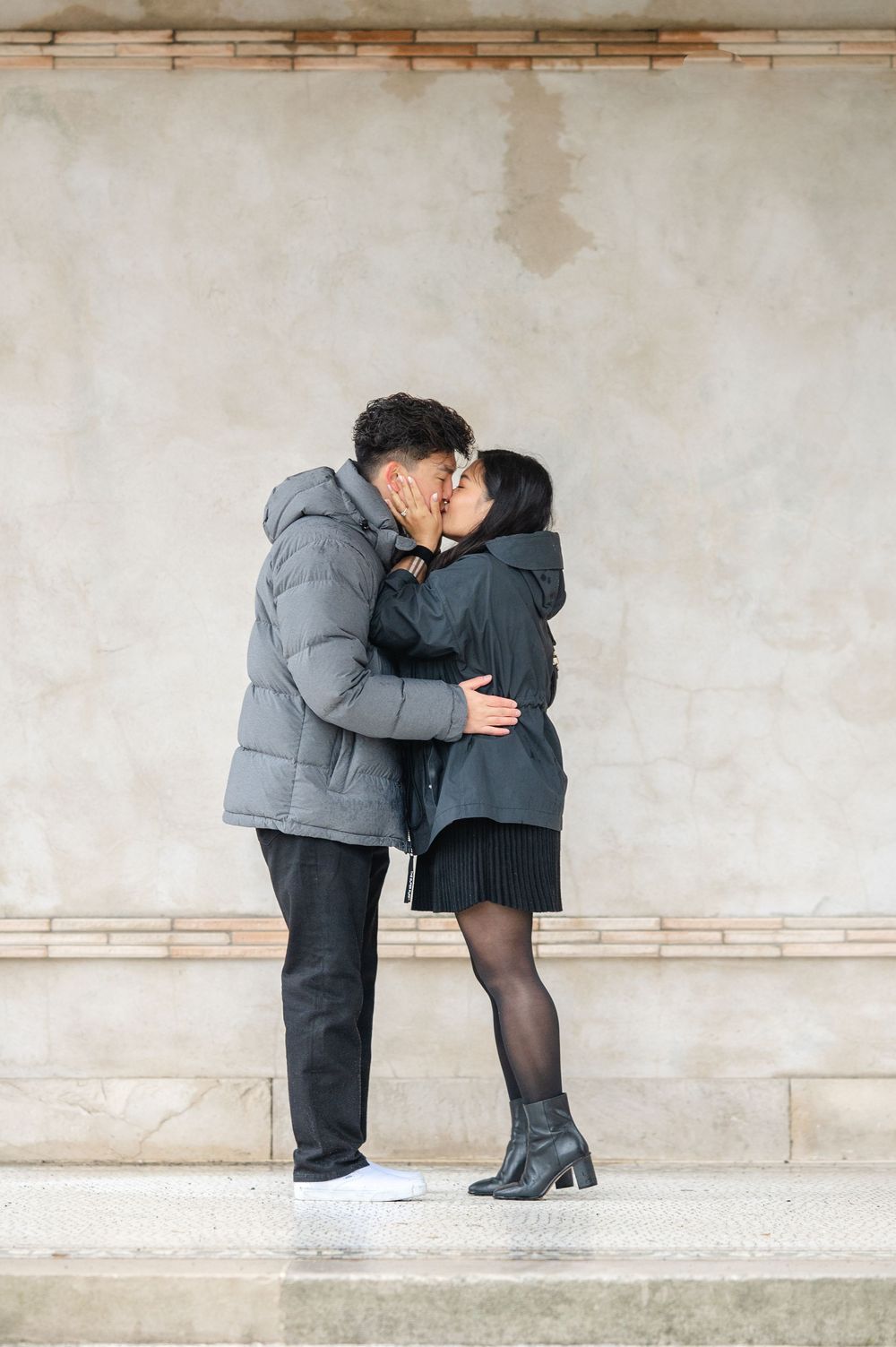 A couple in matching winter coats share an intimate moment while standing against a beige wall after a proposal