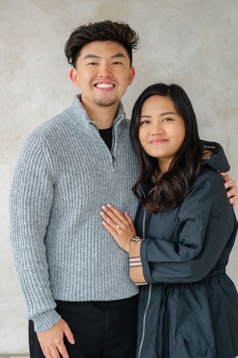 A couple wearing matching gray and navy outfits pose for an engagement photo against a neutral background in Untermyer Gardens