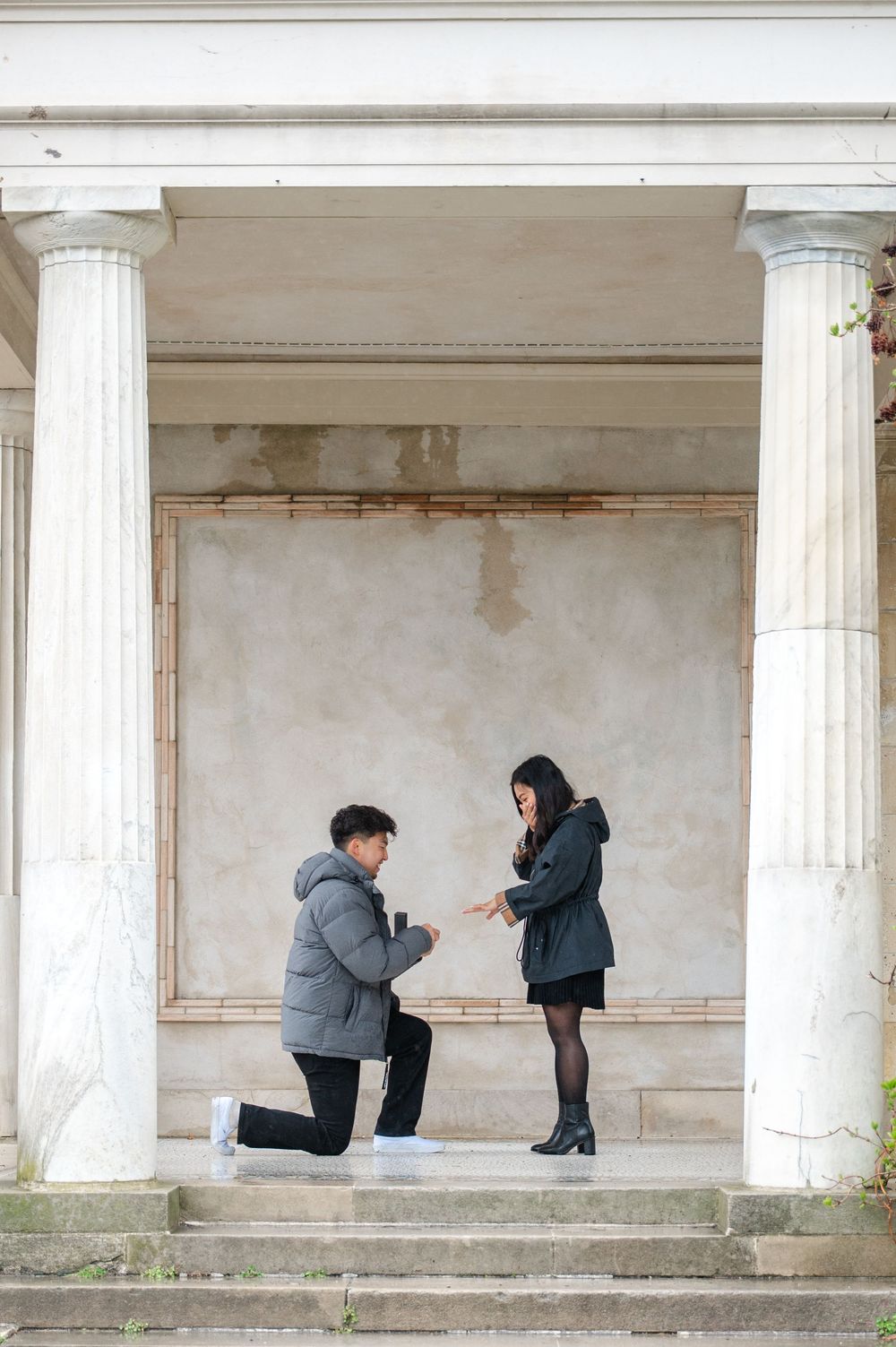 A marriage proposal moment captured between white columns of a classical building at Untermyer Gardens in Westchester, NY outside of NYC