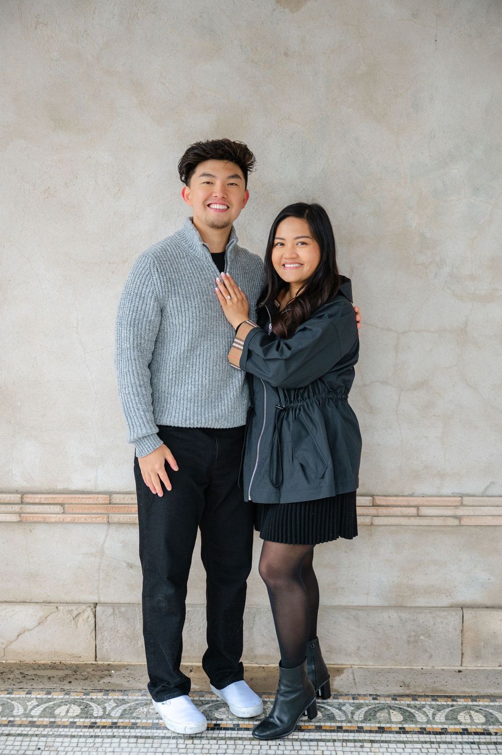 A couple in casual winter outfits pose together against a textured wall with decorative floor tiles in Untermeyer Gardens