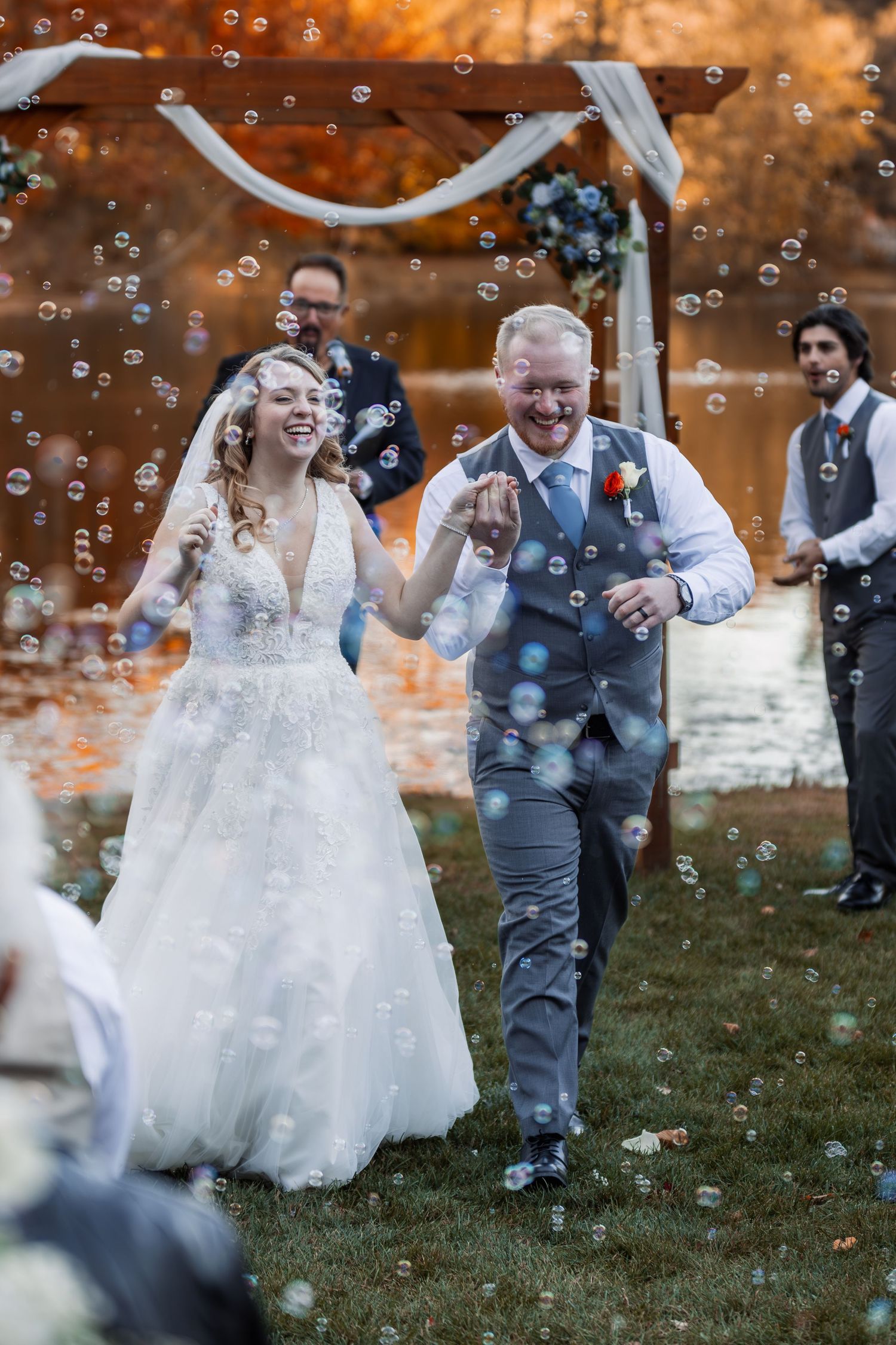 Joyful couple laughs while running through floating bubbles at an autumn outdoor wedding ceremony.