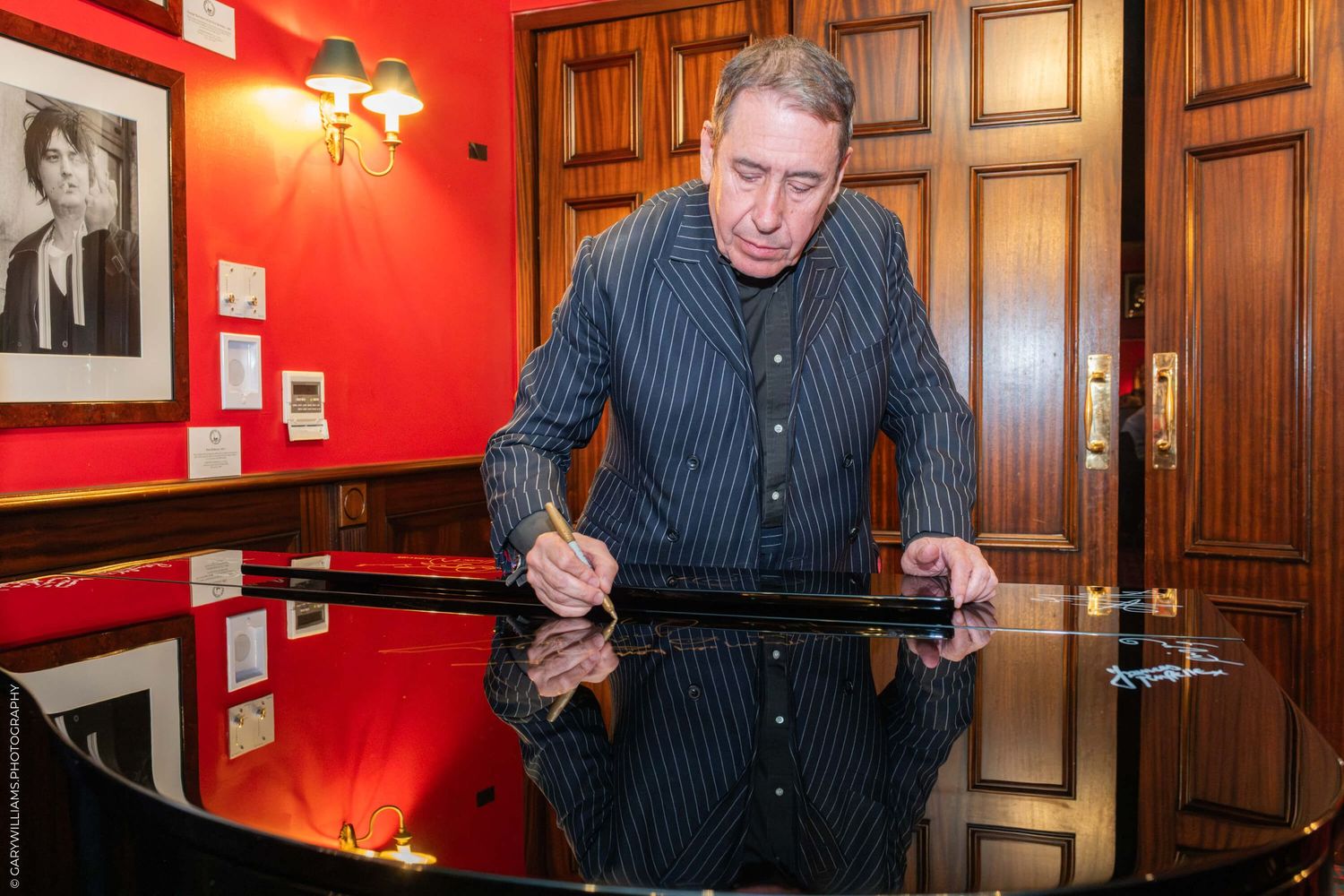 A person in a striped cardigan signs a document at a shiny piano in a room with red walls and wood paneling.