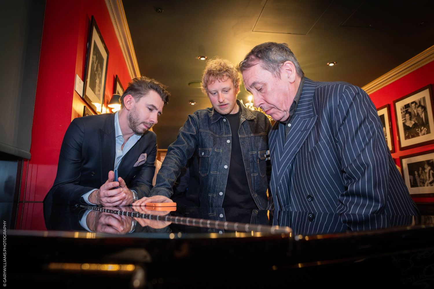 Three people gathered around a piano examining sheet music or documents in a room with red walls.