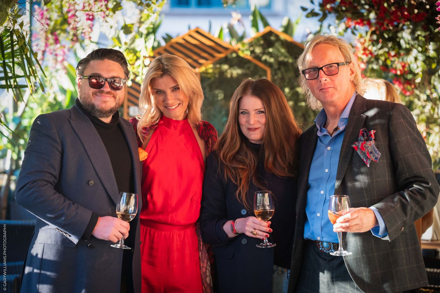 Four people in formal attire standing together holding wine glasses on an outdoor patio with greenery in background.