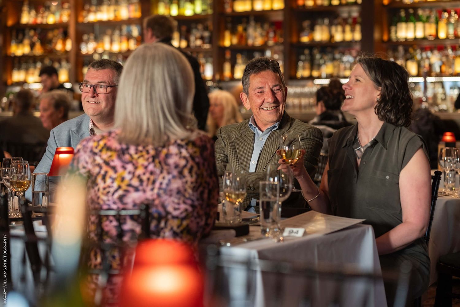 A group of people enjoying drinks and laughing together at a well-stocked bar with ambient lighting.