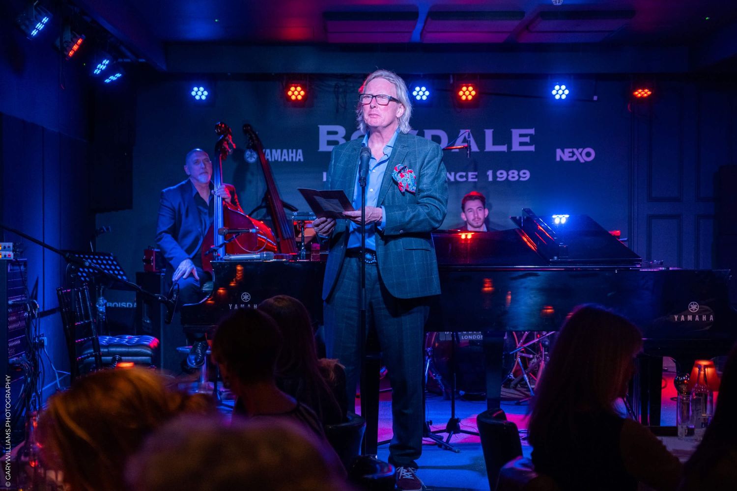 A performer sings on stage at an intimate music venue with blue lighting while a band plays in the background.