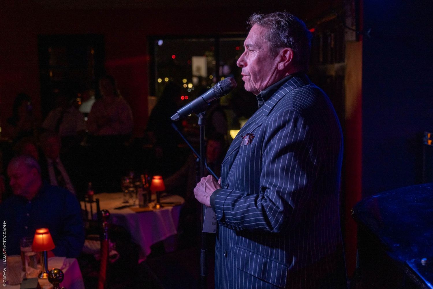 A performer in a pinstriped suit sings at a dimly lit nightclub venue with tables and ambient lighting in the background.