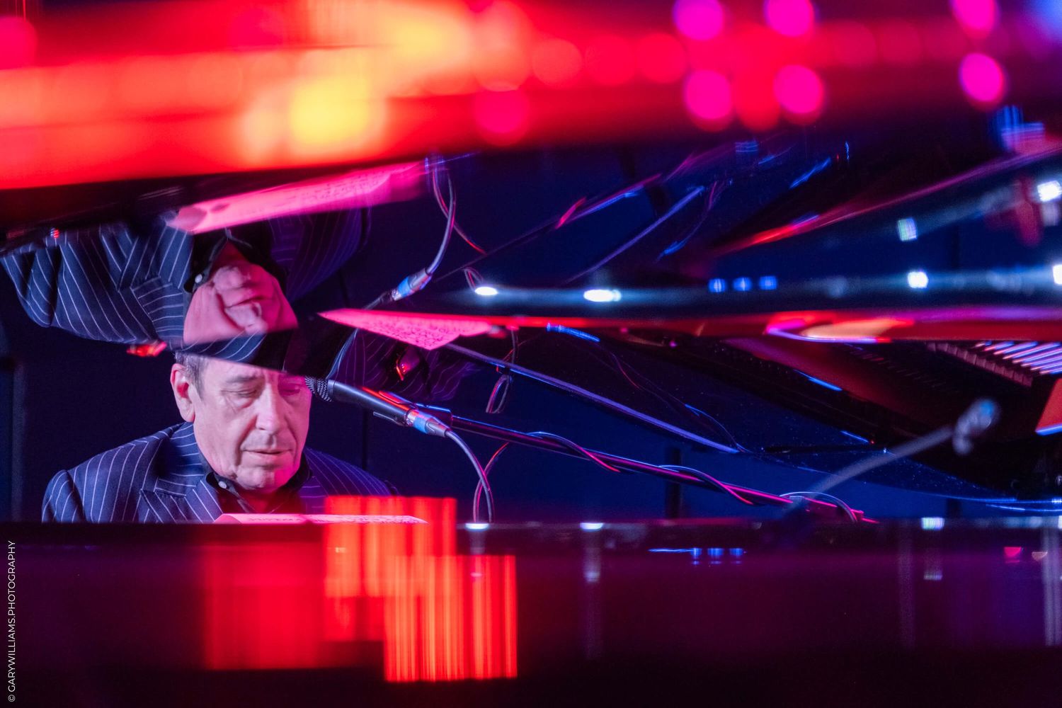Musician performs at piano under dramatic red and blue stage lighting in dark concert setting.