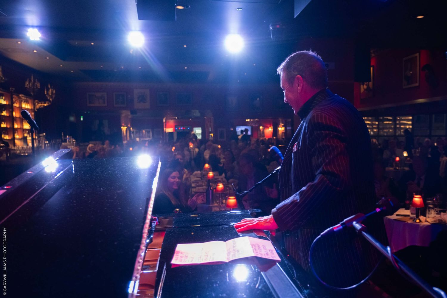 A performer reads from a podium on stage with bright overhead spotlights in a dimly lit venue.