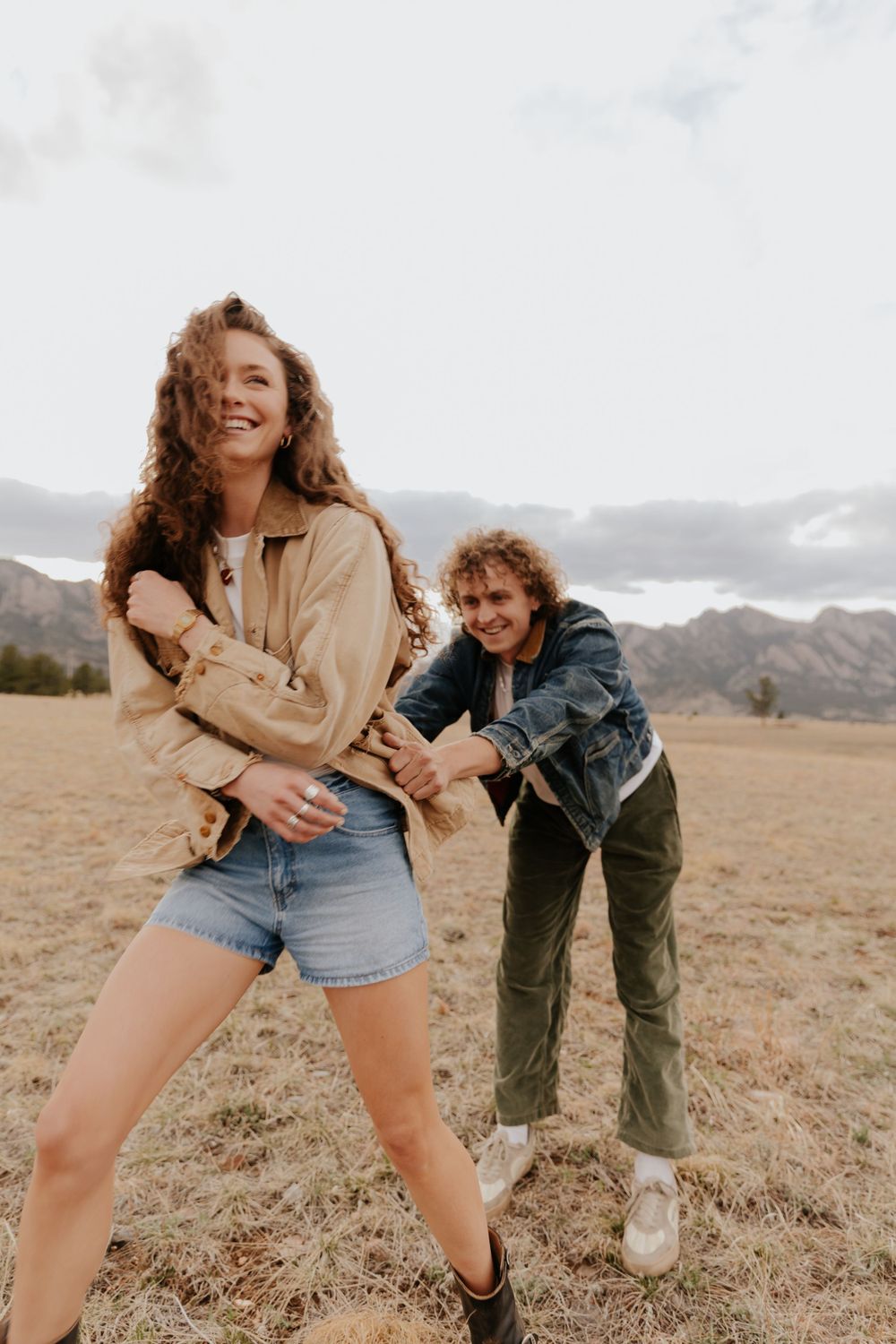 Two people laugh and play together in a field with mountains in the background on an overcast day.