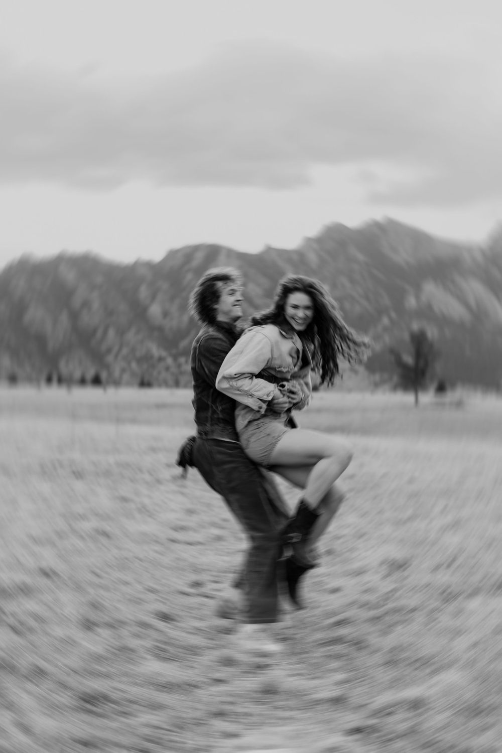 A couple spins and dances together in a field with mountain peaks silhouetted against cloudy skies in black and white.