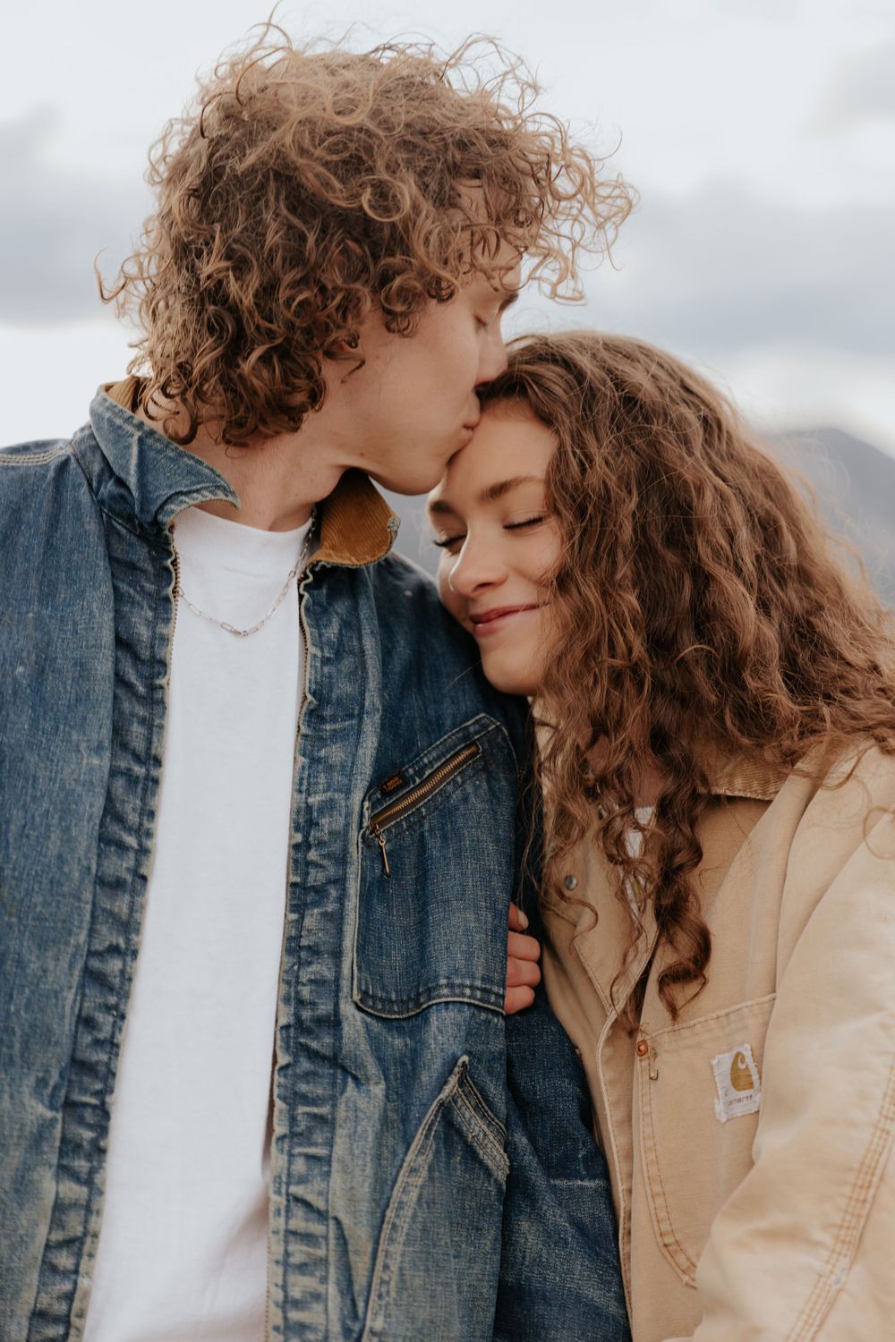 Couple with curly hair sharing an intimate moment outdoors while wearing denim and tan jackets.