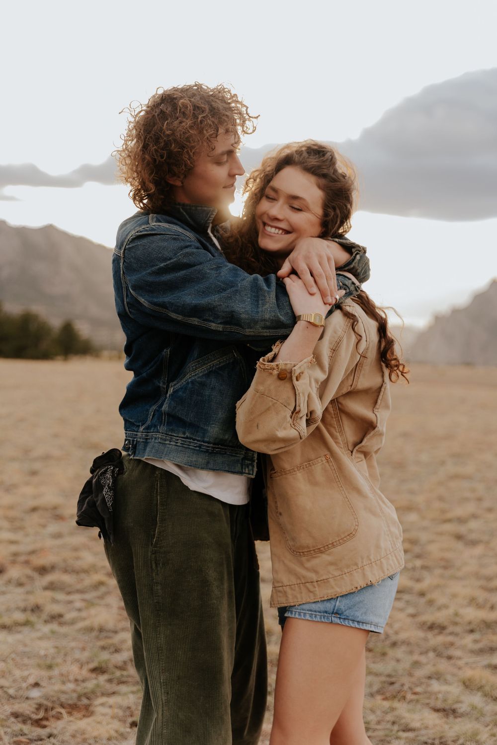 A couple embraces tenderly in a golden field with mountains in the background during sunset.