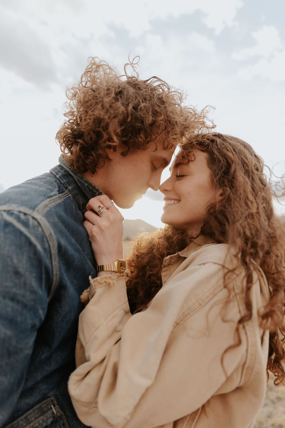 A romantic couple with curly hair embraces outdoors, wearing denim jacket and beige sweater against a cloudy sky.