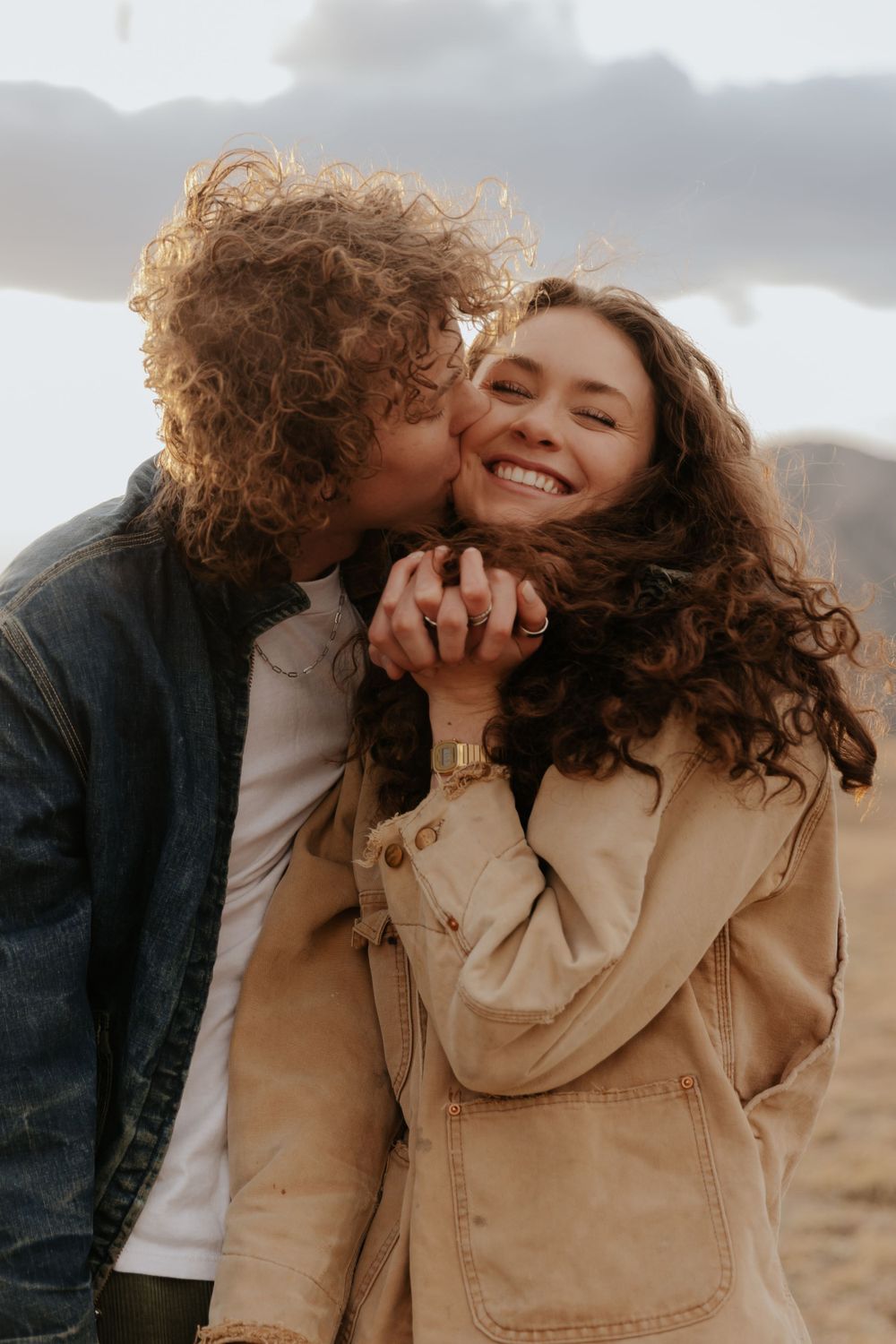 Two people share an intimate outdoor moment together as windswept curly hair frames their joyful expressions in warm lighting.
