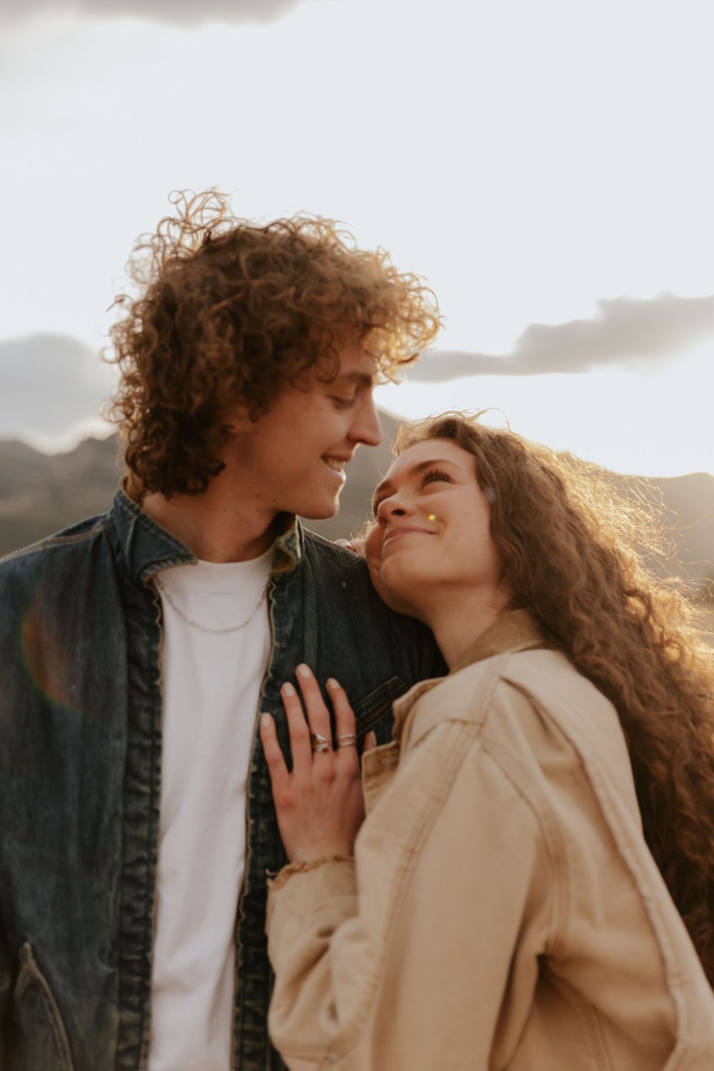A romantic couple sharing an intimate moment outdoors at sunset with soft lighting and curly hair blowing in the breeze.