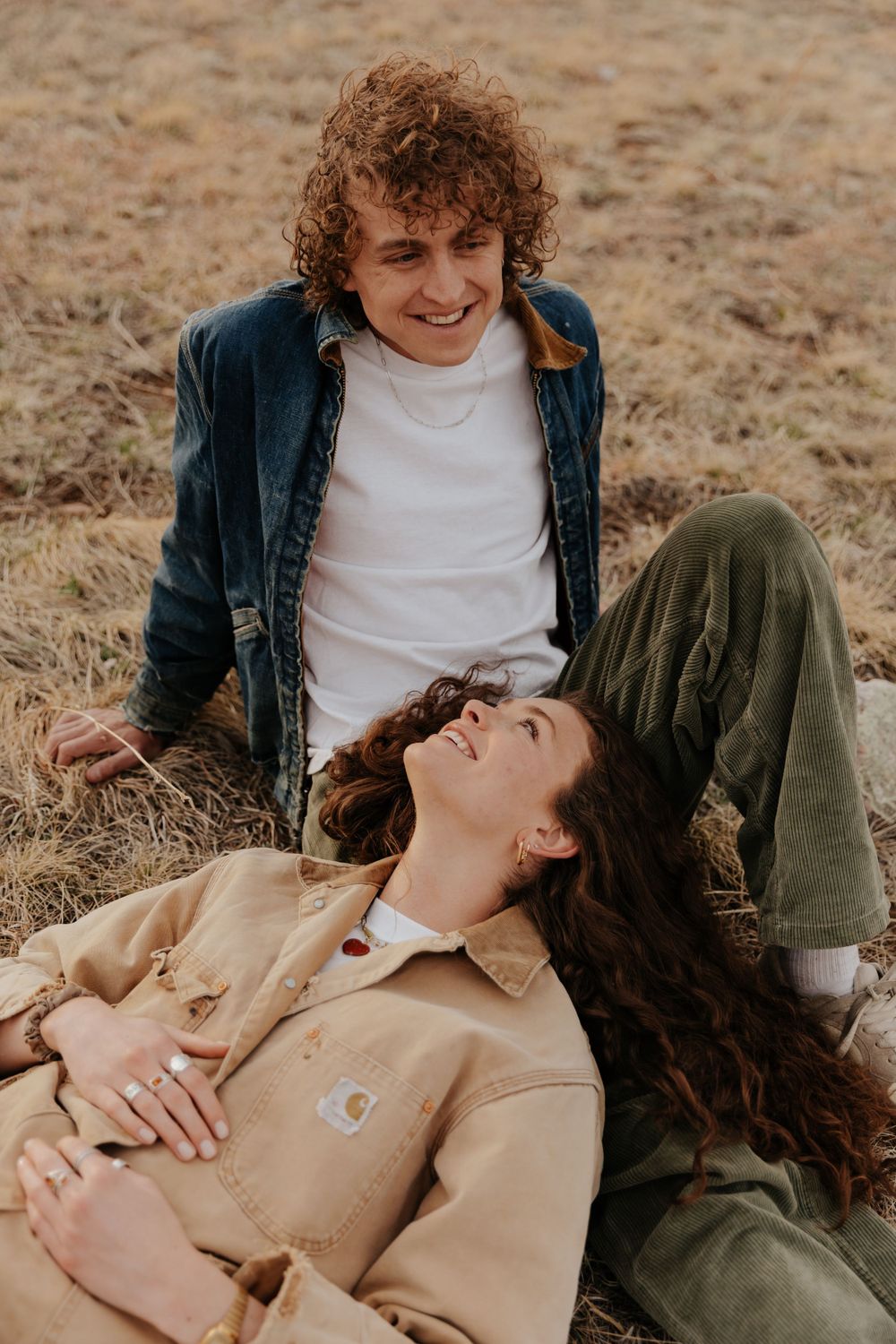 Two people relaxing in a casual autumn setting on dry grass wearing denim and neutral colored clothing.