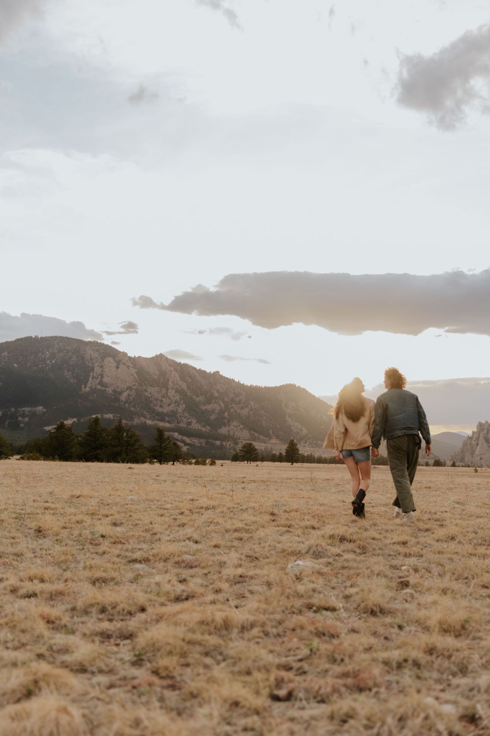 Two people walk together through a grassy field at sunset with mountains in the background.