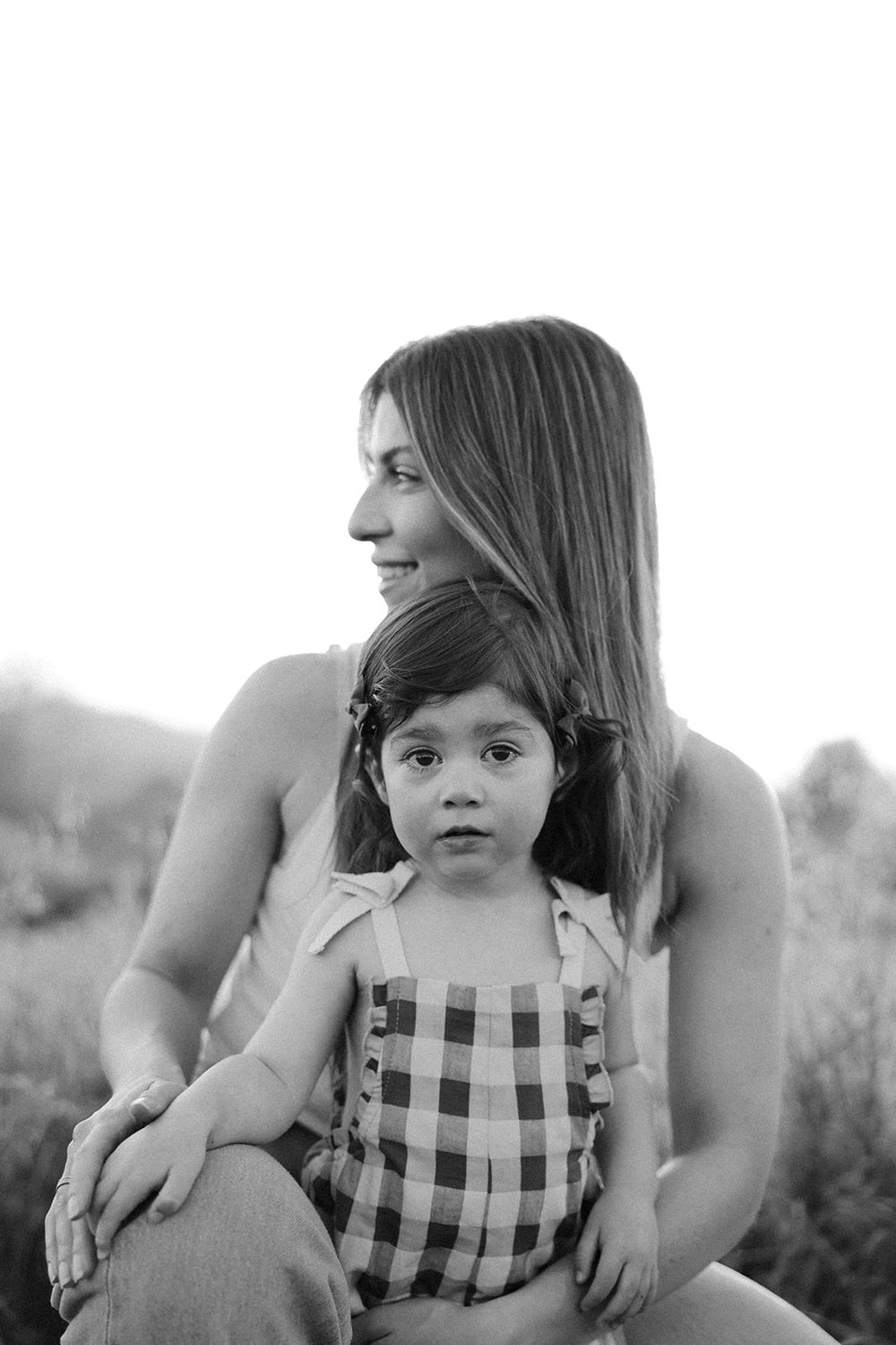 Black and white portrait of parent and child in checkered dress sitting together in sunlit outdoor setting.