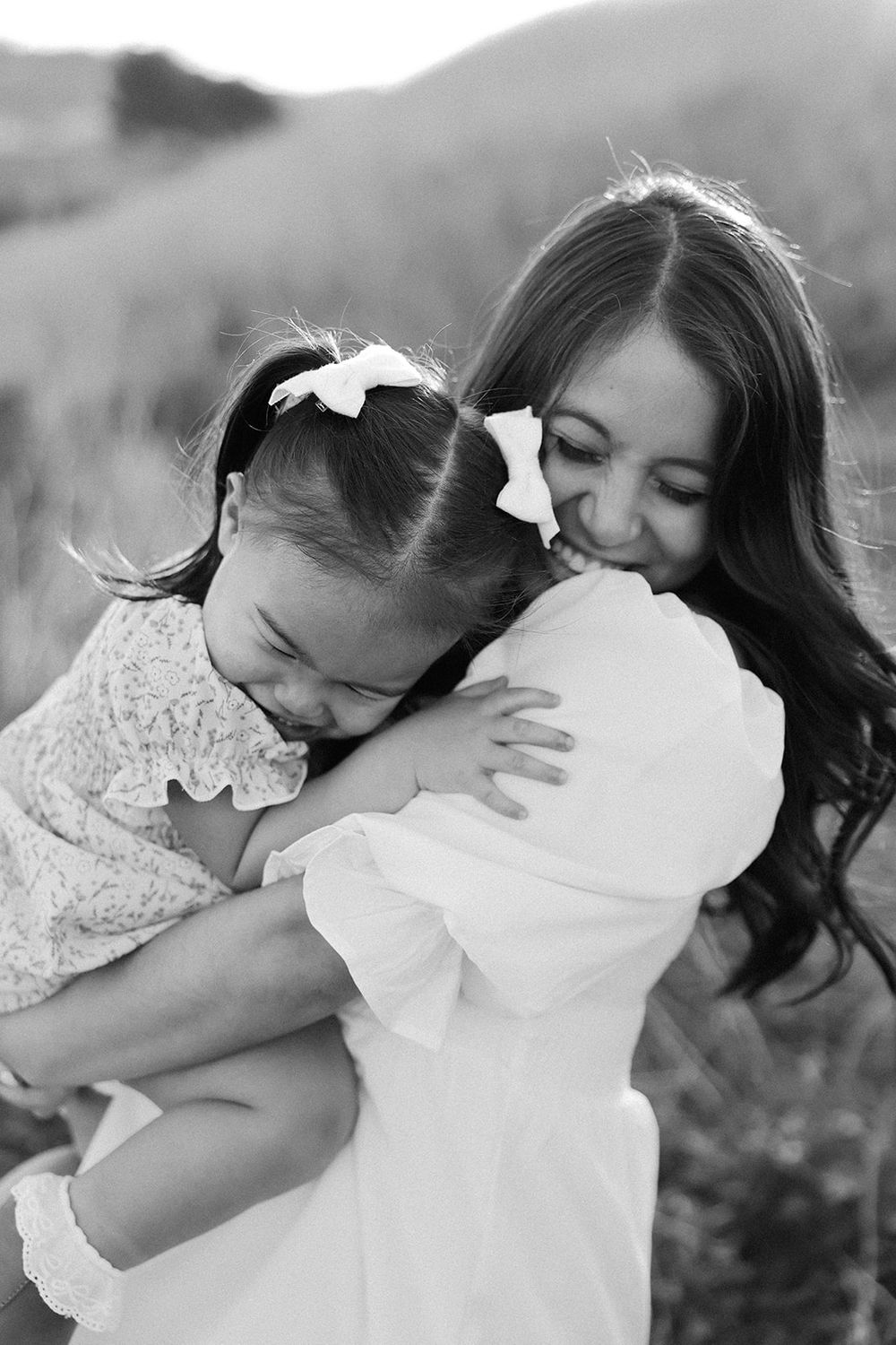 A tender black and white portrait of a mother and child embracing in an outdoor setting, both wearing white dresses.