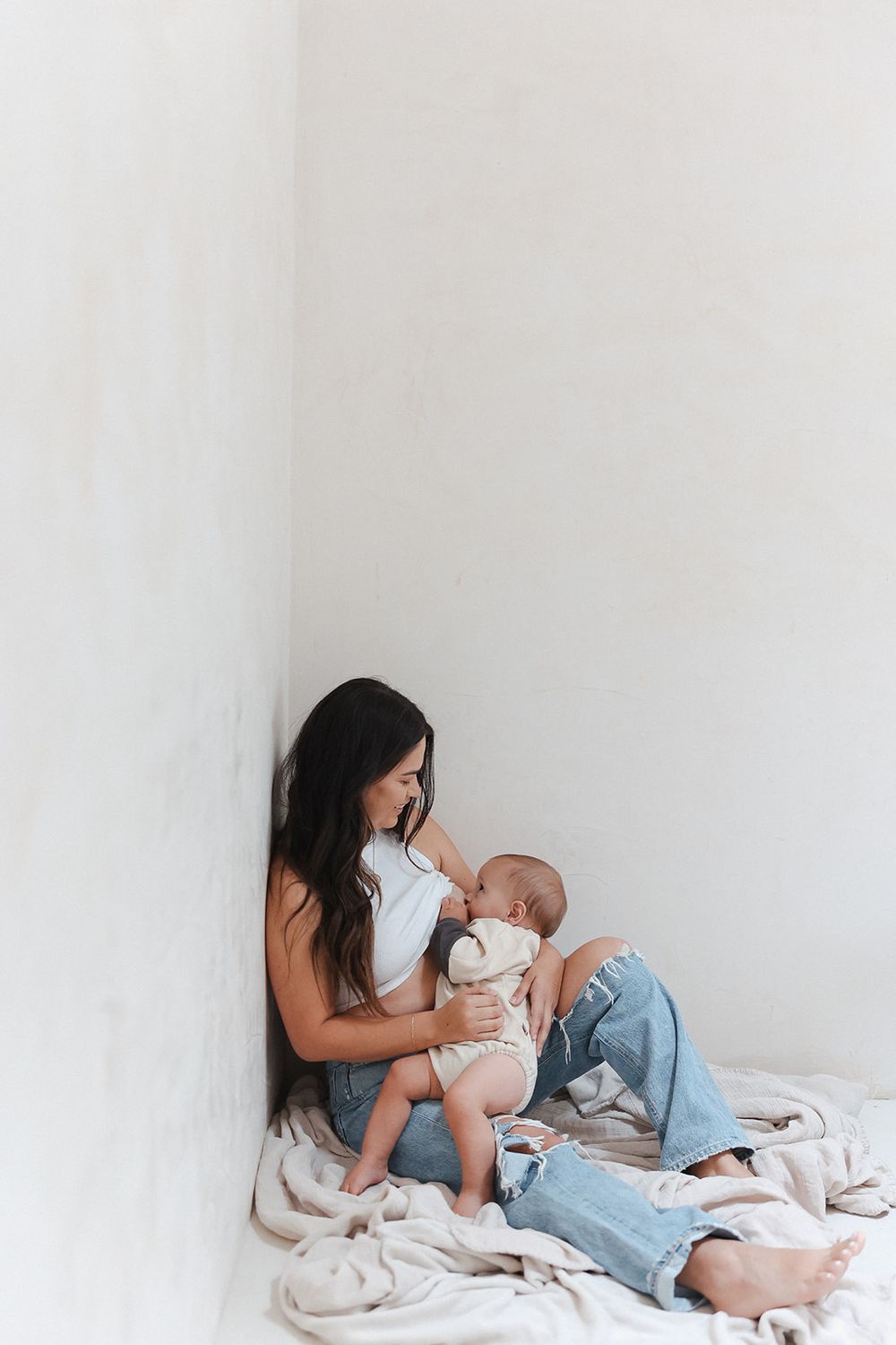 A mother sits on a white floor with ripped jeans while holding and gazing lovingly at her baby against a plain background.