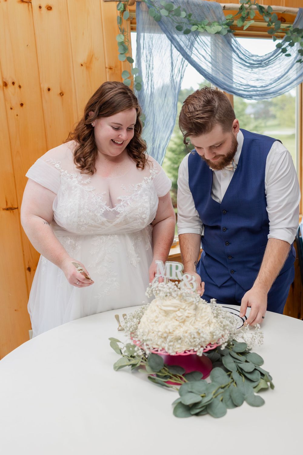 A couple in wedding attire cutting their white decorated cake adorned with greenery at their reception.