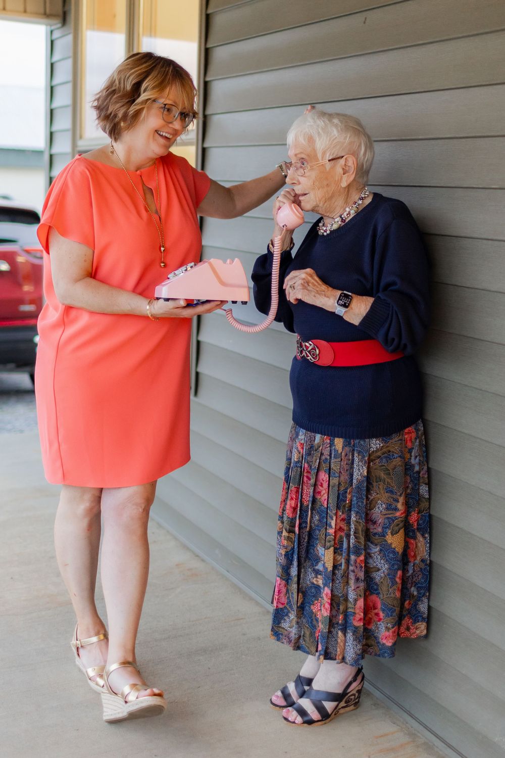 Caregiver in coral dress offers support to elderly woman wearing floral skirt and navy sweater on outdoor porch.