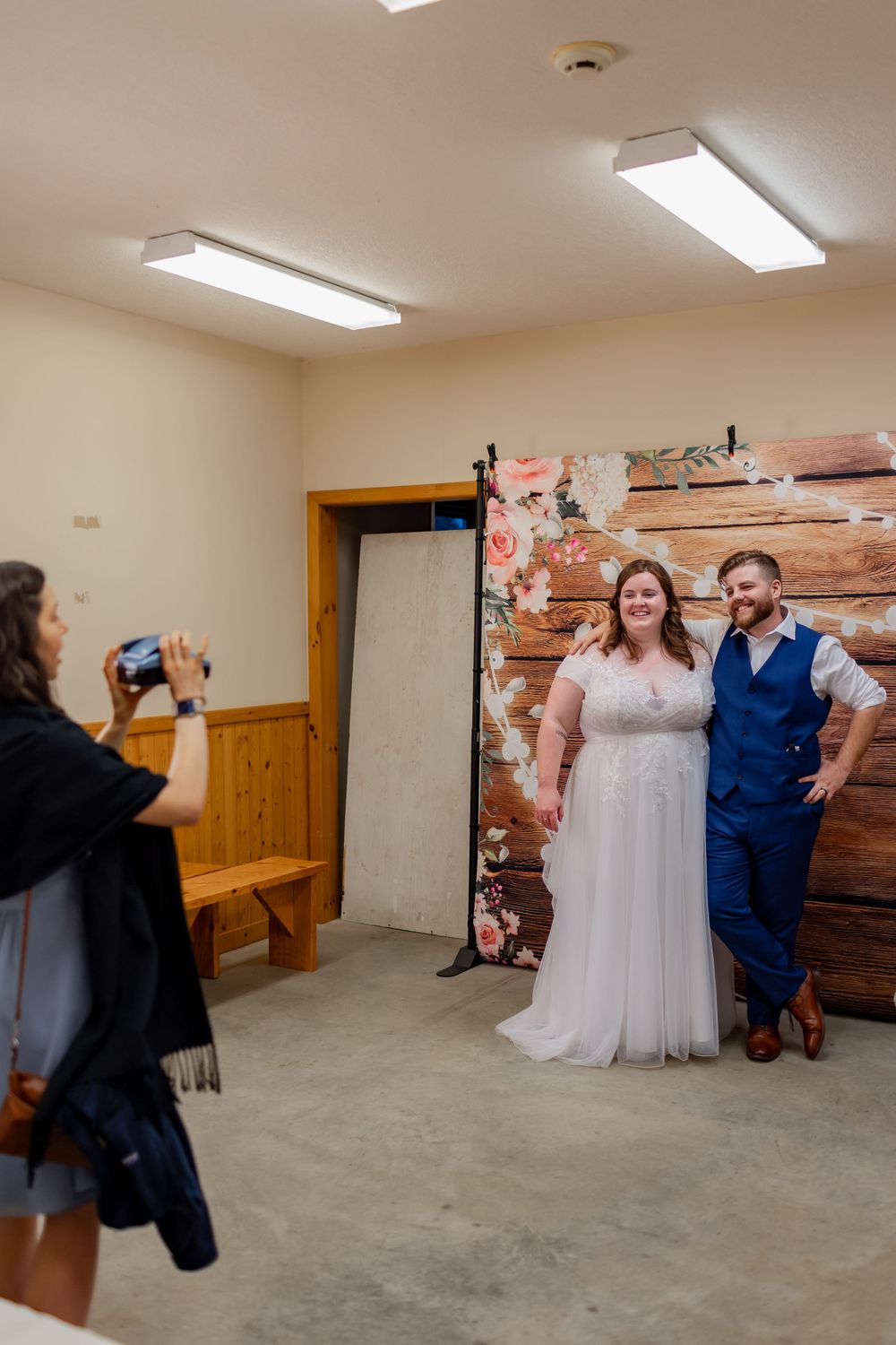 A photographer takes pictures of a couple posing in front of a rustic wooden backdrop in an indoor setting.