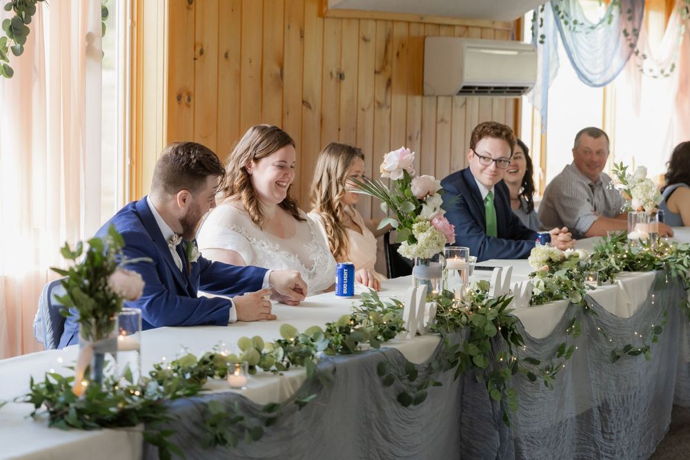 Wedding party gathered at a decorated head table with elegant greenery garland and white floral arrangements.