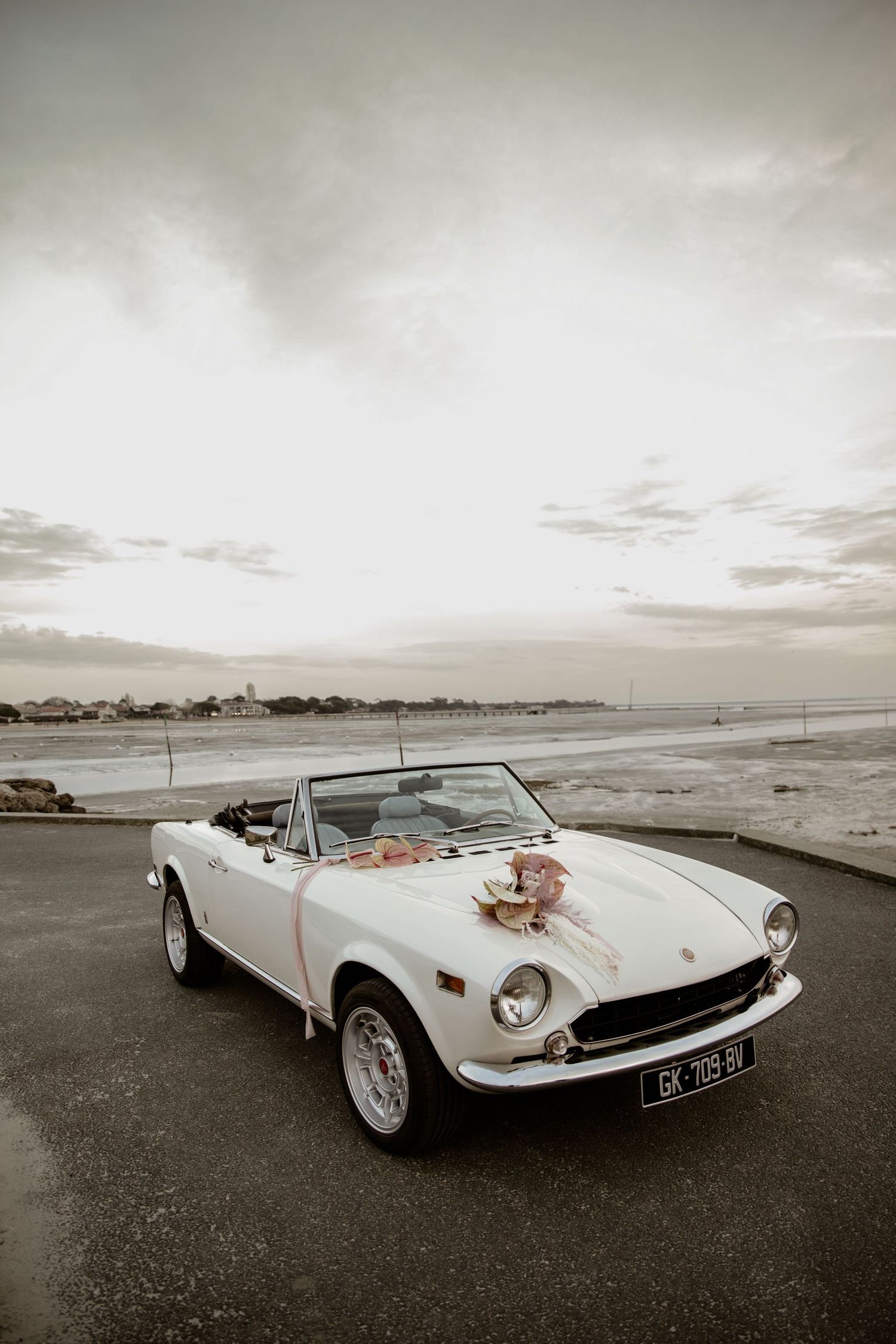 A vintage white Fiat convertible decorated with flowers stands against a coastal sunset backdrop.
