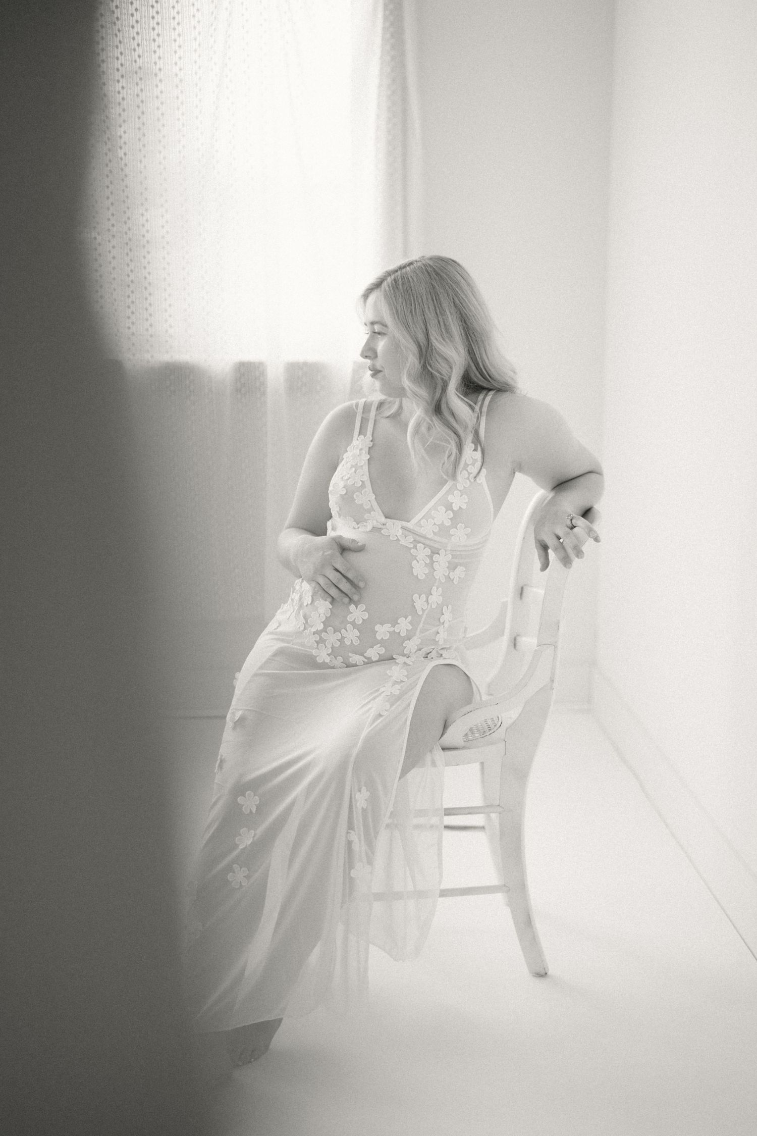 A dreamy black and white boudoir photograph shows a figure in white lingerie seated gracefully on a rustic chair.