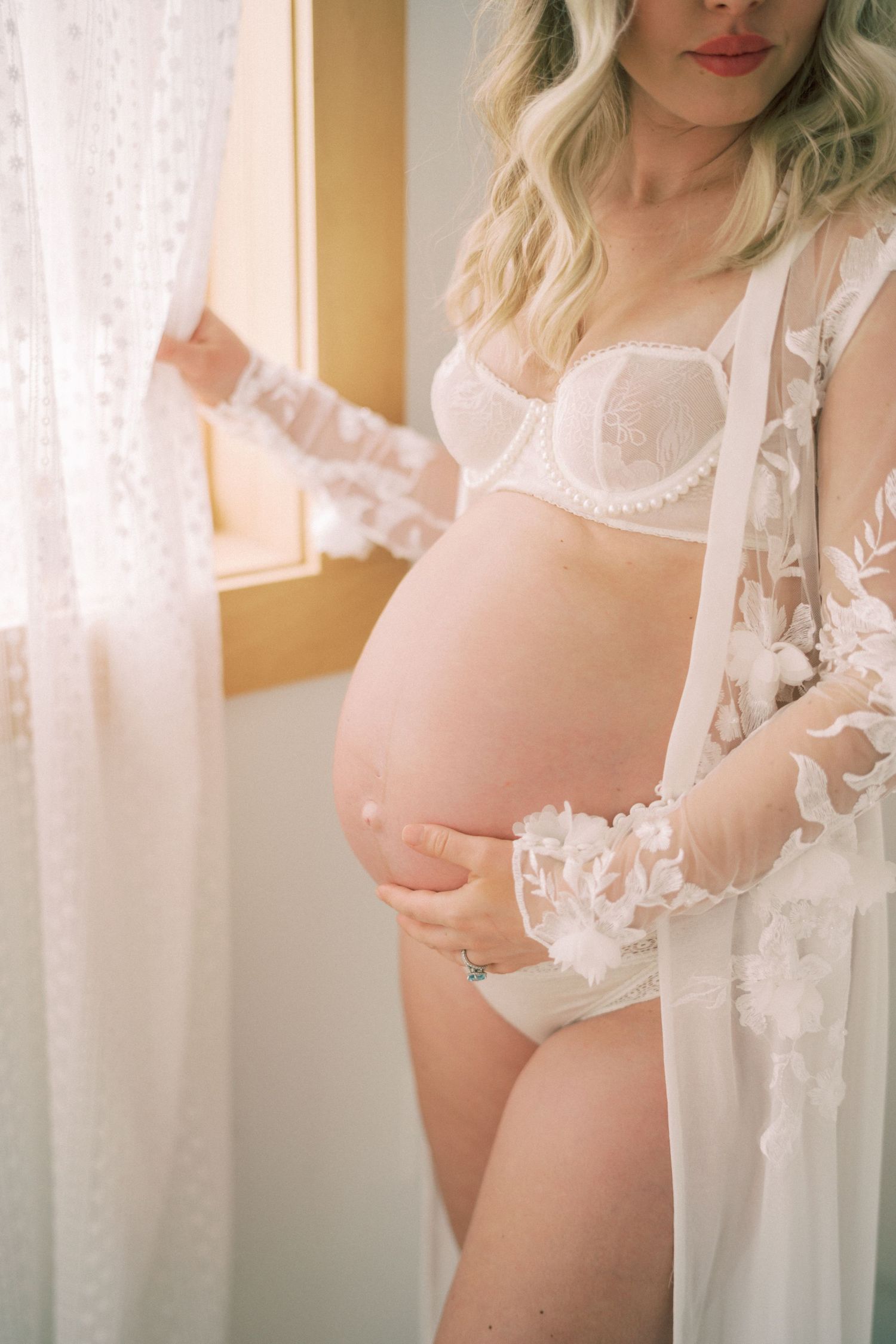 Intimate maternity boudoir photo with lace robe and white lingerie in soft window light.