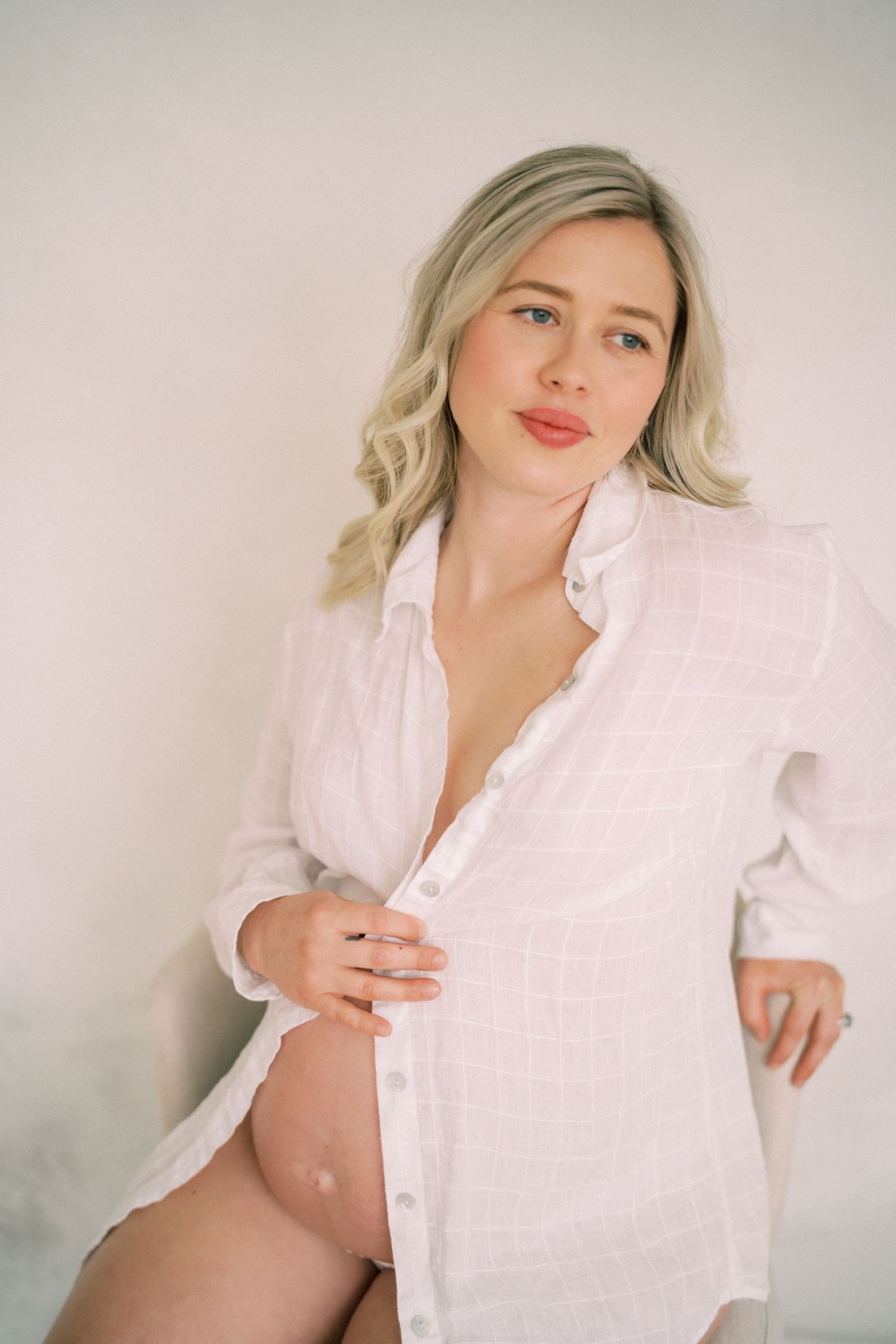 Artistic maternity portrait of a woman wearing an oversized white button-down shirt against a light background.