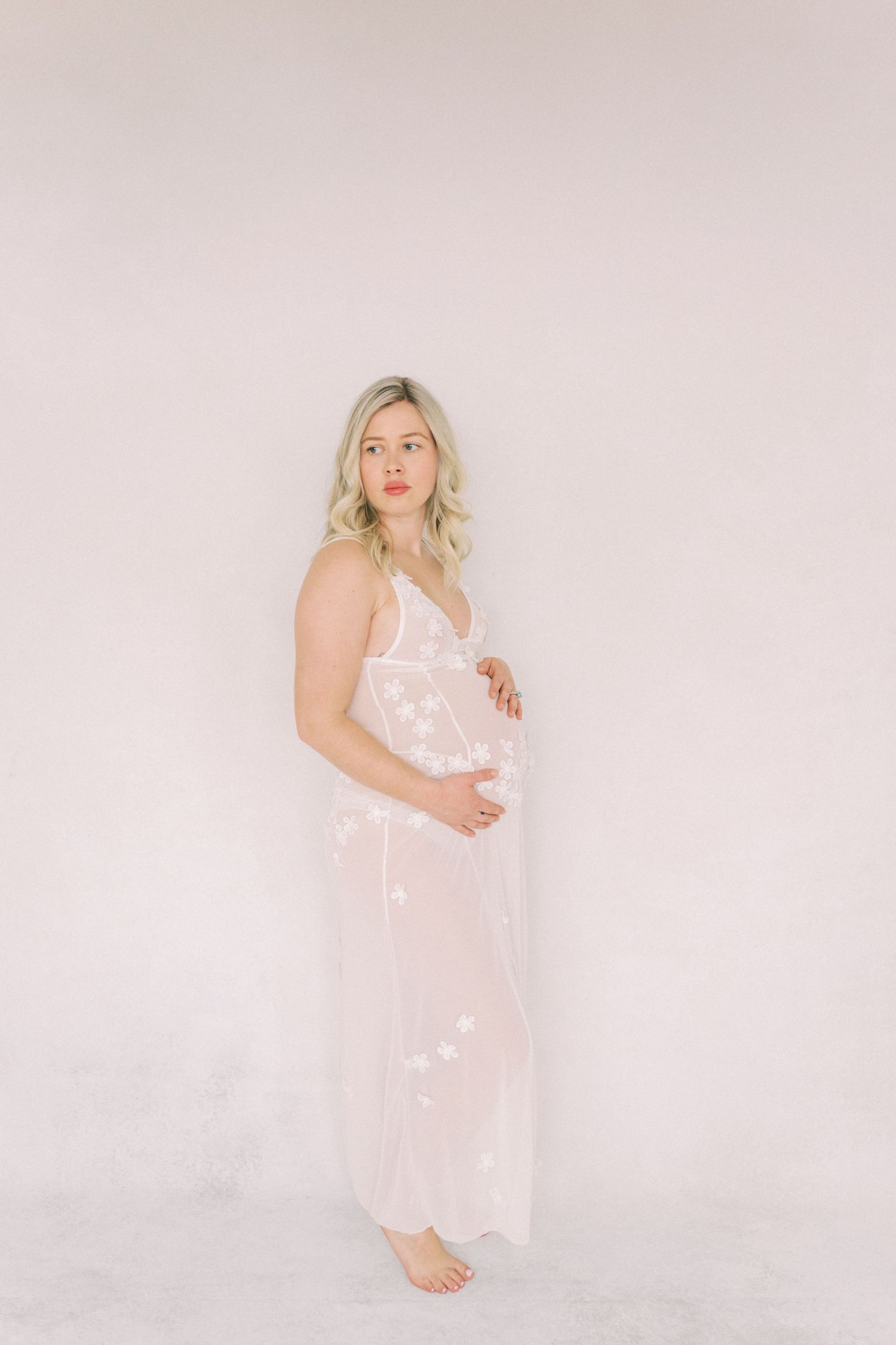 Elegant maternity portrait featuring a blonde woman in a flowing white lace nightgown against a pale pink backdrop.